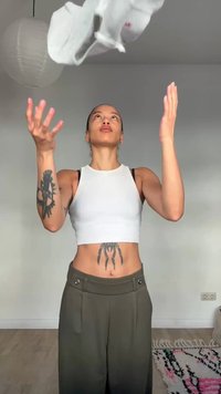 Jongere met tatoeages gooit wit textiel omhoog in een minimalistische kamer met een papieren lantaarn en een patroonrijk vloerkleed.