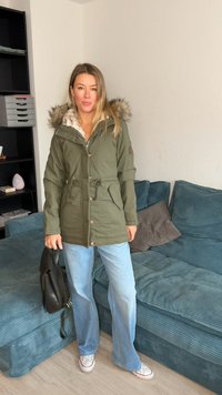 Parka verde oliva com capuz de pelo sintético, cintura com cordão ajustável e fecho de botões. Combinada com jeans de pernas largas azul claro e mala preta.