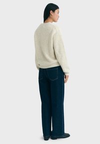 Frau, die seitlich steht, trägt einen gepunkteten cremefarbenen Pullover, dunkelblaue weit geschnittene Jeans und schwarze Schuhe vor einem einfarbigen Hintergrund.