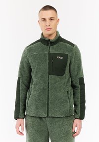 Hombre vestido con chaqueta de forro polar verde oliva y pantalones a juego, de pie de frente, con bolsillos con cremallera en el pecho y los laterales, contra un fondo blanco.