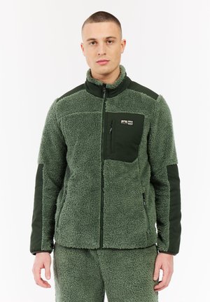 Man in een olijfgroene fleecejack en bijpassende broek, recht naar voren kijkend, met ritszakken op de borst en zijkanten, staand tegen een witte achtergrond.