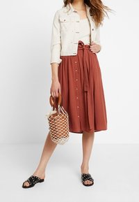 Veste en jean clair, crème ; débardeur côtelé, beige ; jupe midi couleur rouille avec boutons ; sandales noires à clous ; sac tissé.
