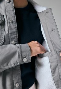 Grå denimjakke med blød hvid fleecefór, trykknapper og en inderlomme med en stofsløjfe til sikker opbevaring.