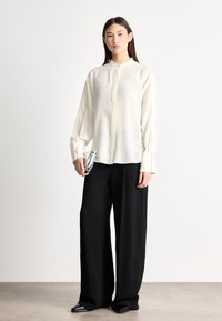 Fehér selyem ing mandzsettás gallérral, gombos elülső résszel és hosszú ujjakkal, széles fekete nadrággal és fekete cipővel párosítva. Ezüst borítéktáskát tart a kezében.