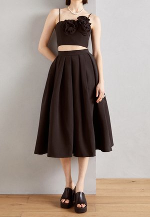 Femme portant un crop top noir sans manches avec des détails floraux et une jupe midi noire plissée taille haute avec des sandales plateformes noires.