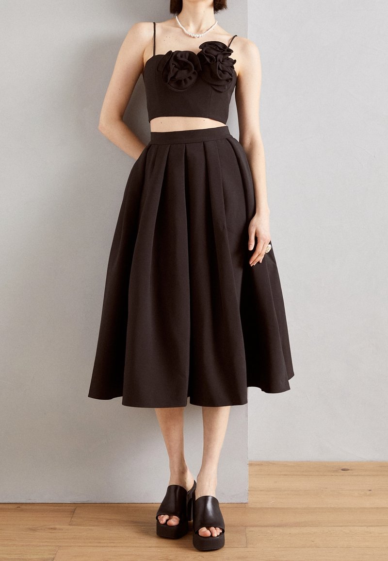 Femme portant un crop top noir sans manches avec des détails floraux et une jupe midi noire plissée taille haute avec des sandales plateformes noires.