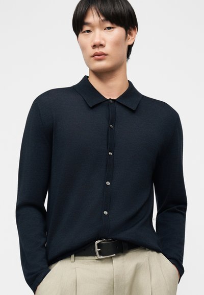 Νέος άνδρας που φοράει μαύρο κουμπωτό ζακέτα με γιακά και μπεζ πιεττέ παντελόνι με μαύρη ζώνη, χέρια στις τσέπες.