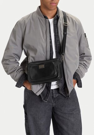 Hombre con chaqueta bomber gris claro, camisa vaquera negra, vaqueros oscuros y bolso de cuero negro cruzado con bolsillo frontal y parche con logo.