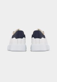 Ζευγάρι λευκά sneakers χαμηλού προφίλ με παχιά λευκή σόλα και σκούρες ναυτικές λεπτομέρειες στη φτέρνα, εμφανιζόμενα από την πίσω πλευρά σε απλό φόντο.