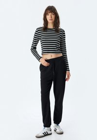 Schwarz-weiß gestreiftes, kurzes Langarmoberteil mit figurbetontem Design; kombiniert mit lockeren, schwarzen Sweatpants und weißen Sportschuhen.