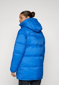 Blauwe pufferjack met een capuchon, voorzien van een gestikte ontwerp, elatische manchetten en een gladde, glanzende afwerking. De achterkant is iets langer.