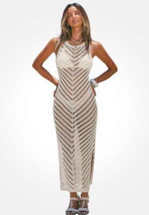 Kobieta w bezrękawnym, beżowym, dzianinowym maksisukienki z wzorem i rozcięciem po boku, nosząca sandały z koralików i biżuterię z muszli, pozująca z zamkniętymi oczami.