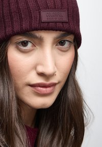 Gorro de punto burdeos con textura acanalada y un parche rectangular que dice "LADOS COLORIDOS DE LA VIDA". Primer plano de un rostro que lleva el gorro.