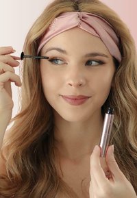 Tube de mascara avec une finition rose doré brillante tenu dans une main, appliquant le produit avec un applicateur noir dans l'autre main. Arrière-plan rose doux.
