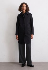 Frau mit lockigem Haar, die einen schwarzen knöpfen Mantel, weite graue Hosen und schwarze Slipper trägt, steht vor einer weißen strukturierten Wand.