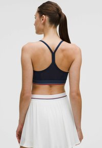 Granatowy stanik sportowy z designem typu racerback, kontrastowym wykończeniem i gładką fakturą. W zestawie z plisowaną białą spódnicą.