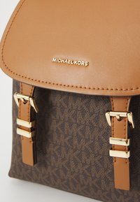 Sac marron à motif logo avec un rabat en cuir beige, des garnitures en or et des sangles ajustables, présentant un design épuré et structuré.