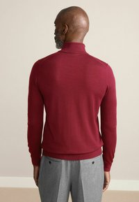 Pulover burgundy cu guler înalt, cu o textură moale, design ajustat și manșete cu nervuri. Asortat cu pantaloni gri plisati, care au buzunare în spate.