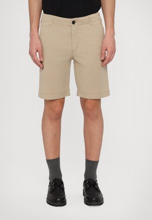 Beige cotton shorts with a straight cut, rolled hem, and front pockets, paired with gray socks and black shoes.