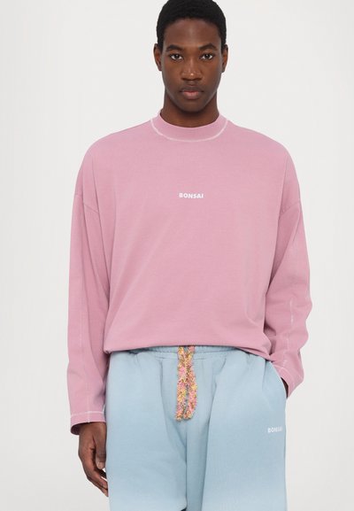 Rozā garām piedurknēm krekls ar "BONSAI" logotipu, apvienots ar gaiši ziliem sporta biksēm, kas piedāvā daudzkrāsainu auklu. Kokvilnas materiāls, brīvs piegriezums.