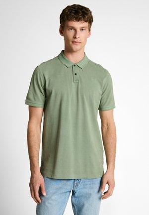 Young man with short curly hair wearing a light green polo shirt and light blue jeans, standing against a plain white background.