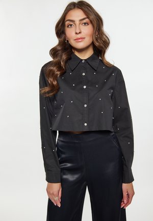 Zwarte cropped blouse met lange mouwen, voorzien van een knoopsluiting aan de voorkant en verfraaid met kleine, glanzende accenten. Gecombineerd met glanzende zwarte broek.