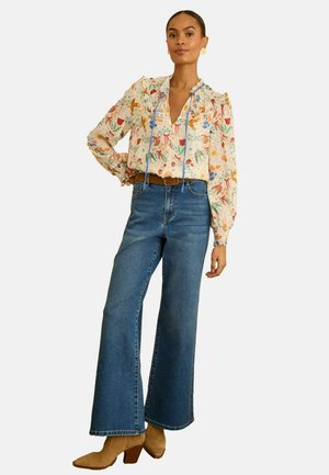 Sieviete, valkājot ziedotu garroku blūzi, ieliktu augsta vidukļa platām zilām džinsu biksēm ar brūnu jostu un bēšām papēžu zābakiem.