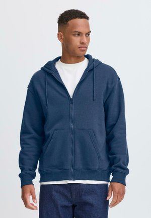 Felpa con zip color navy, con cordoncini, tasche frontali e polsini a costine. Indossata sopra una maglietta bianca, esposta su uno sfondo chiaro.