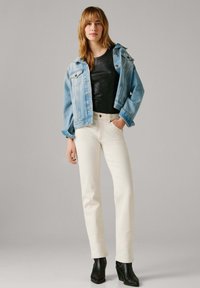 Γυναίκα που φοράει ανοιχτό μπλε τζιν σακάκι πάνω από μαύρο τοπ, λευκά παντελόνια με ψηλή μέση και μαύρα μποτάκια αστραγάλου, στέκεται με το ένα χέρι στην τσέπη.