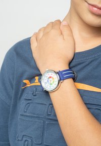 Montre-bracelet en silicone bleu avec un cadran circulaire arborant des marqueurs d'heure colorés et des chiffres. Portée au poignet d'un enfant, aux côtés d'une chemise bleue.