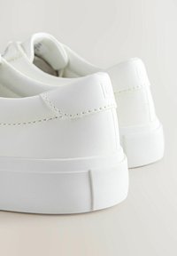 O pereche de adidași albi, cu un design curat, cusături vizibile și talpa groasă din cauciuc, pe un fundal neutru.