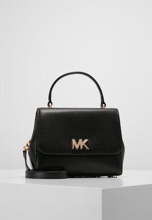 Schwarze Lederhandtasche mit Tragegriff oben, goldenem "MK"-Logo auf der vorderen Klappe und abnehmbarem Schulterriemen, präsentiert auf einem weißen Podest.