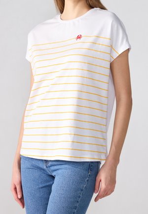 Kobieta w białym krótkim T-shircie z cienkimi żółtymi poziomymi paskami i małym czerwonym detalem kraba przy kołnierzu, zestawiona z niebieskimi dżinsami.