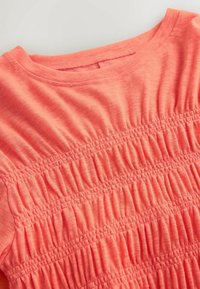 Tissu rose corail avec des bandes élastiques horizontales froncées créant une texture plissée au niveau de l’encolure et du haut de la poitrine du vêtement.