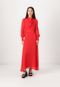 Piros sifon maxi ruha magas nyakkal, megkötő részlettel, hosszú puffos ujjal és karcsú derékkal. Fekete platform szandállal párosítva.