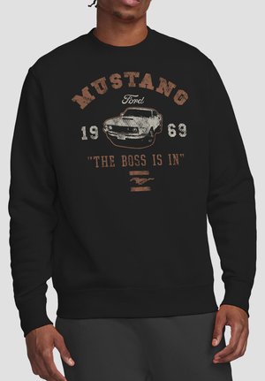 Person wearing black sweatshirt with vintage white and brown Mustang car graphic, "Ford 1963," and text "THE BOSS IS IN."