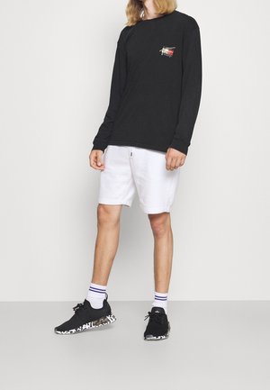 Zwart shirt met lange mouwen en een logo, witte korte broek en zwarte sneakers met een patroon op de zool, gecombineerd met witte sokken met strepen.