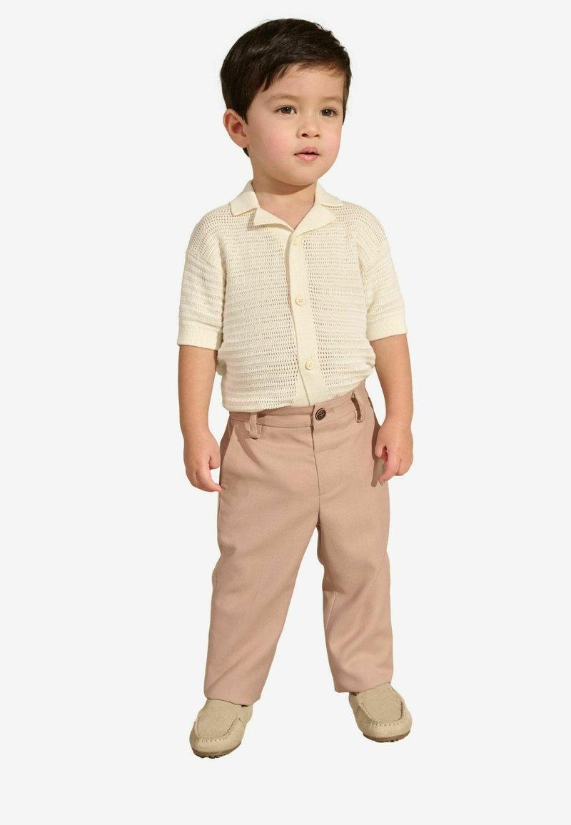 Hellbeige kurzärmliges Strickshirt mit Kragen, kombiniert mit hellrosa Hosen und beigen Mokassins. Einfaches Knopfdetail am Shirt.