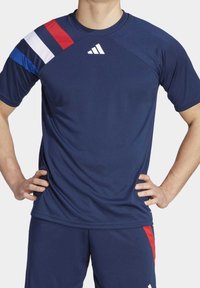 Muž v námořnickém modrém sportovním tričku a šortkách se červenými, bílými a modrými pruhy na rameni a logem Adidas na hrudi.
