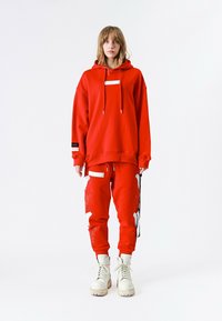 Persona con una sudadera roja de gran tamaño y pantalones joggers a juego con parches blancos, combinados con botas blancas con cordones, de pie sobre un fondo blanco.