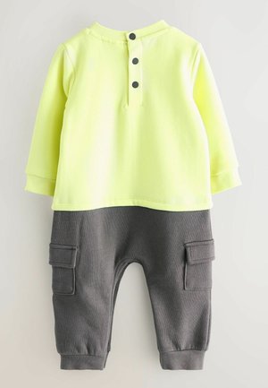 Kollane kapuutsiga särk, mille seljal on kolm musta nuppu, koos hallide cargo-pükstega, millel on küljetaskud ja ribatud püksisääred.