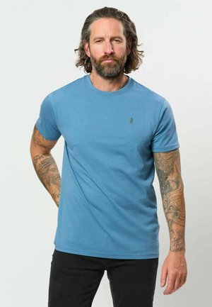 Homme avec barbe et tatouages porte un t-shirt bleu à manches courtes et un pantalon noir, debout devant un fond clair uni.