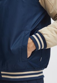 Tamsiai mėlynas bomber tipo švarkas su smėlio spalvos rankovėmis, guma apdailos rankovėse su tamsiai mėlynomis ir kreminėmis juostelėmis bei šone integruota kišenė.