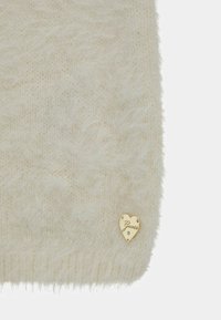 Krämfärgad stickad hatt med mjukt, texturerat tyg och rullad kant, med en guldfärgad hjärtformad loggobricka med "Guess" präntad.
