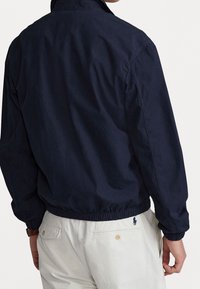 Marineblaue, leichtgewichtige Jacke mit elastischem Saum, sichtbarer Naht und hohem Kragen. Getragen über hellen Hosen mit Logo-Detail.