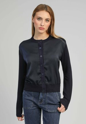 Jeune femme aux cheveux blonds lisses, portant un cardigan noir boutonné et un jean bleu, tournée vers l'avant sur un fond uni.