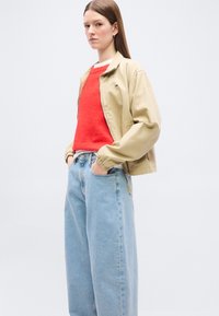 Γυναίκα με ίσια καστανή τρίχα, που φοράει μπεζ σακάκι, λαμπερό κόκκινο πουλόβερ και ανοιχτό μπλε τζιν παντελόνι με φαρδιά πόδια, με τα χέρια στις τσέπες, σε απλό φόντο.