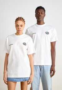 T-shirts de algodão brancas com mangas curtas apresentam o texto "Em Férias" e um gráfico colorido "WORLD TOUR", usados por dois modelos.