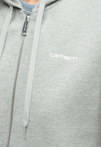Ανοιχτό γκρι φούτερ με φερμουάρ από μαλακό ύφασμα. Διαθέτει αση μένιο φερμουάρ και κεντητό λογότυπο "carhartt" σε λευκό χρώμα στην αριστερή πλευρά.