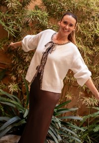 Maglione bianco lavorato a maglia con un fiocco in stampa leopardo al collo, abbinato a una gonna marrone aderente. Circondato da piante verdi.